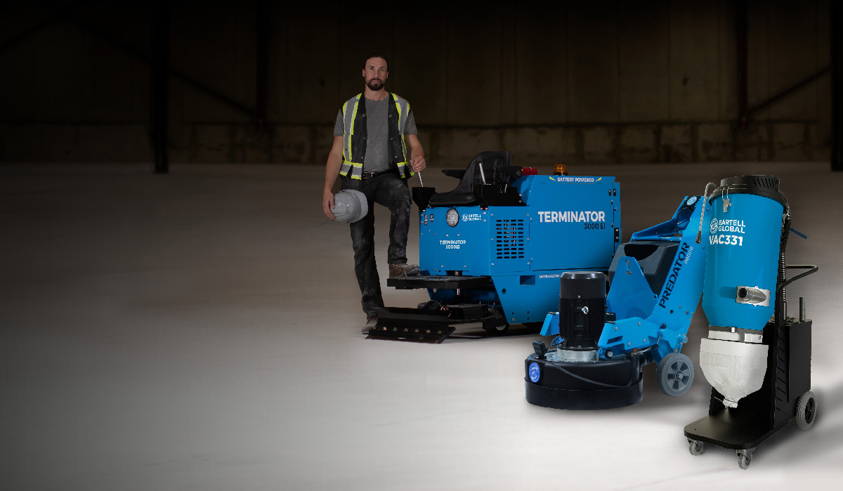 21310-WebSlider-HIRE-final Worker in a safety vest stands beside blue floor grinding machines in a large empty warehouse or hangar setting.